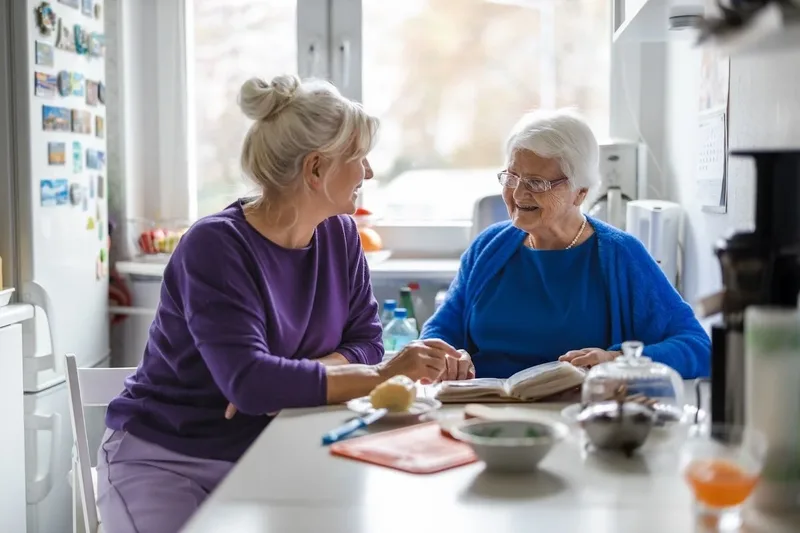 Pflegerin und Seniorin lachen gemeinsam beim Gespräch in der Küche – Seniorenbetreuung