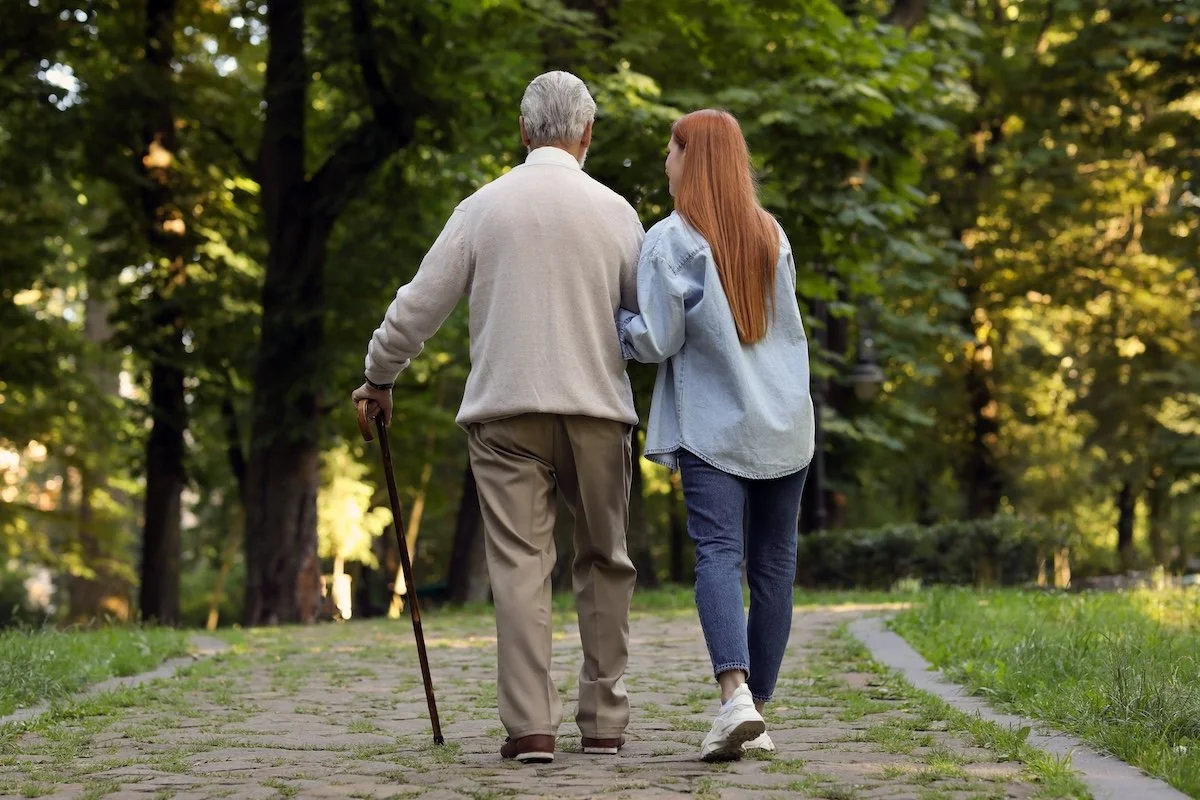 Junge Frau begleitet ihren älteren Vater beim Spaziergang im Park – Pflegende Angehörige