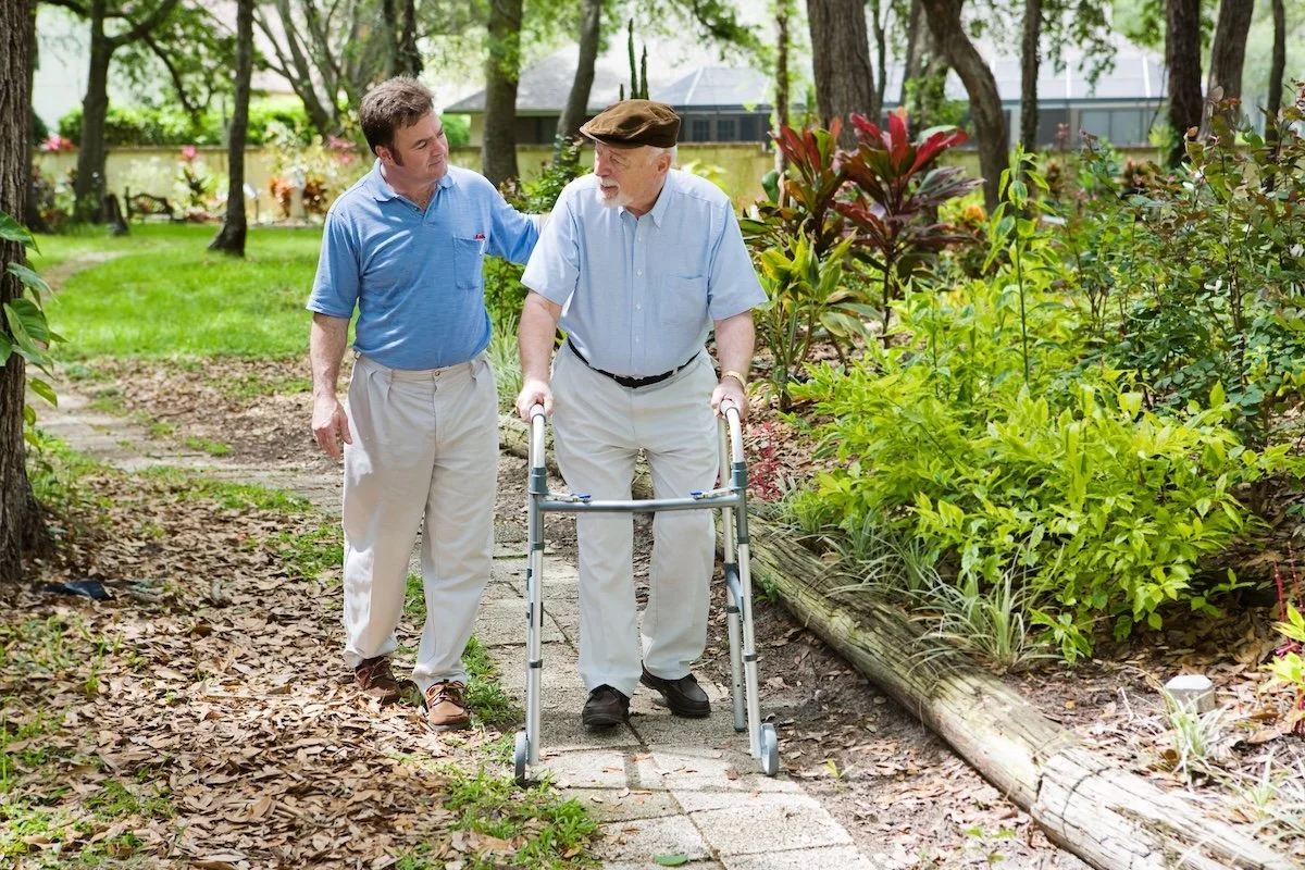 Betreuer begleitet Senior mit Rollator beim Spaziergang im Garten – Ambulante Pflegedienste